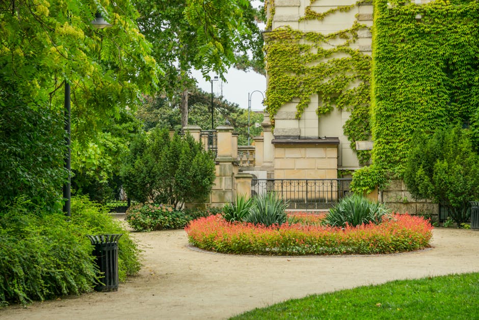 A small landscaped garden area featuring a circular flower bed edged with red and green foliage plants, situated on a tidy gravel pathway. To the left, lush green trees and bushes provide shade and na